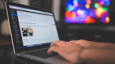 Hands typing on a laptop with a blog post visible, cozy indoor setting with colorful screen in background.